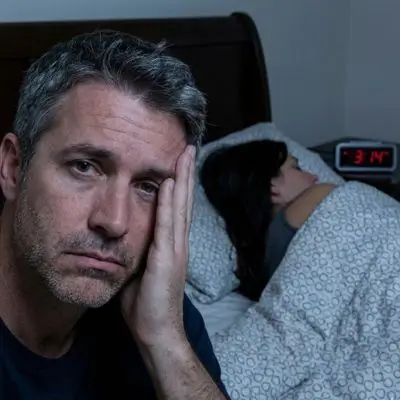 Primo piano di un uomo di 52 anni sveglio a letto di notte con espressione frustrata e triste, mentre la moglie dorme di schiena accanto a lui